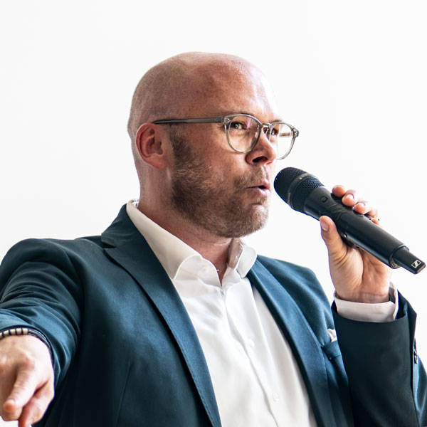 Glatzköpfiger Mann mit Brille spricht in ein Handmikrofon.